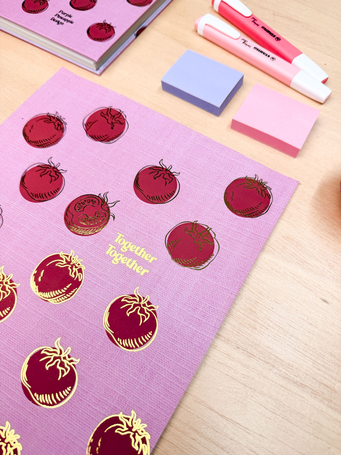 Caderno e folhas decoradas com tomates vermelhos e texto dourado, blocos de notas rosa e roxo e marcadores numa mesa de madeira.