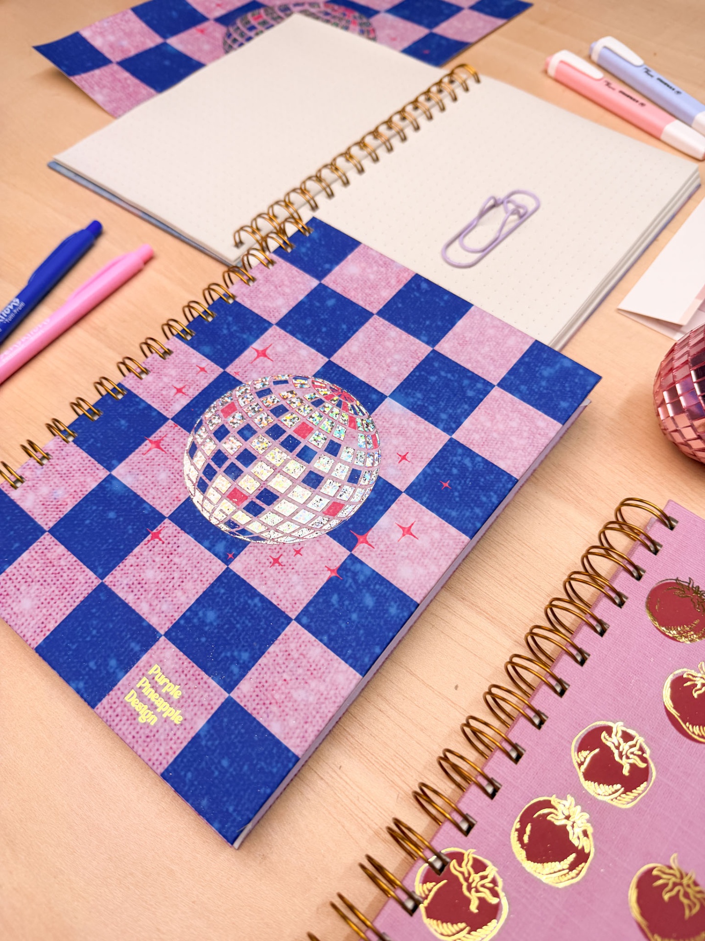 Cadernos espiral decorados com padrões e texto Purple Pineapple Design sobre secretária de madeira com canetas e clips.