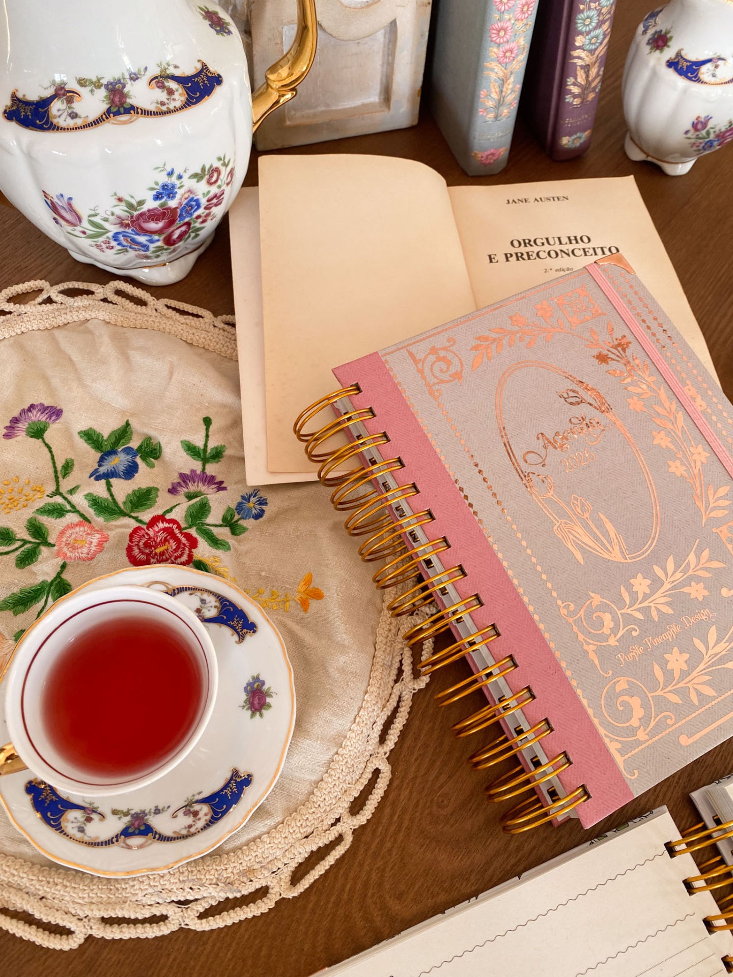 Agenda rosa 2026, chávena com chá, jarra decorativa, livro aberto e toalha bordada sobre mesa de madeira