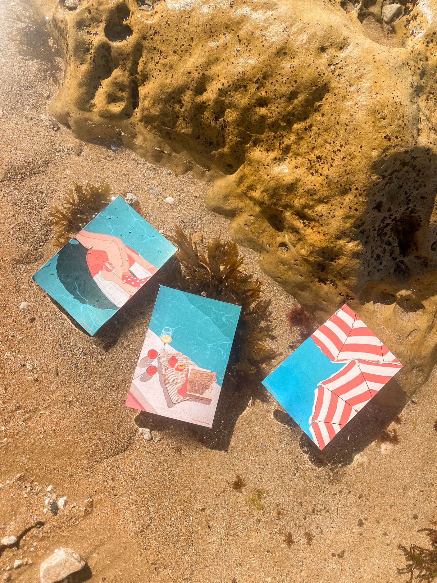 Três postais coloridos de praia na areia molhada junto a uma rocha com algas