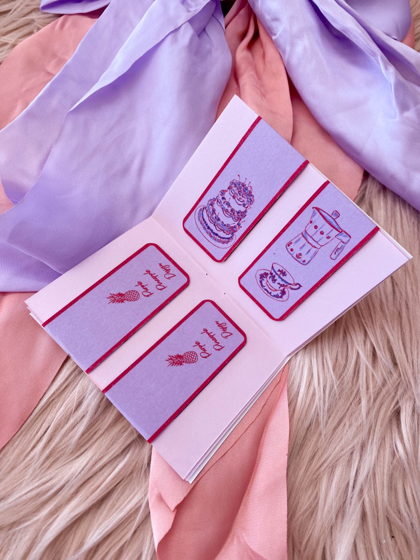 Marcadores de livros lilás com ilustrações vermelhas sobre tecidos rosa e lilás.