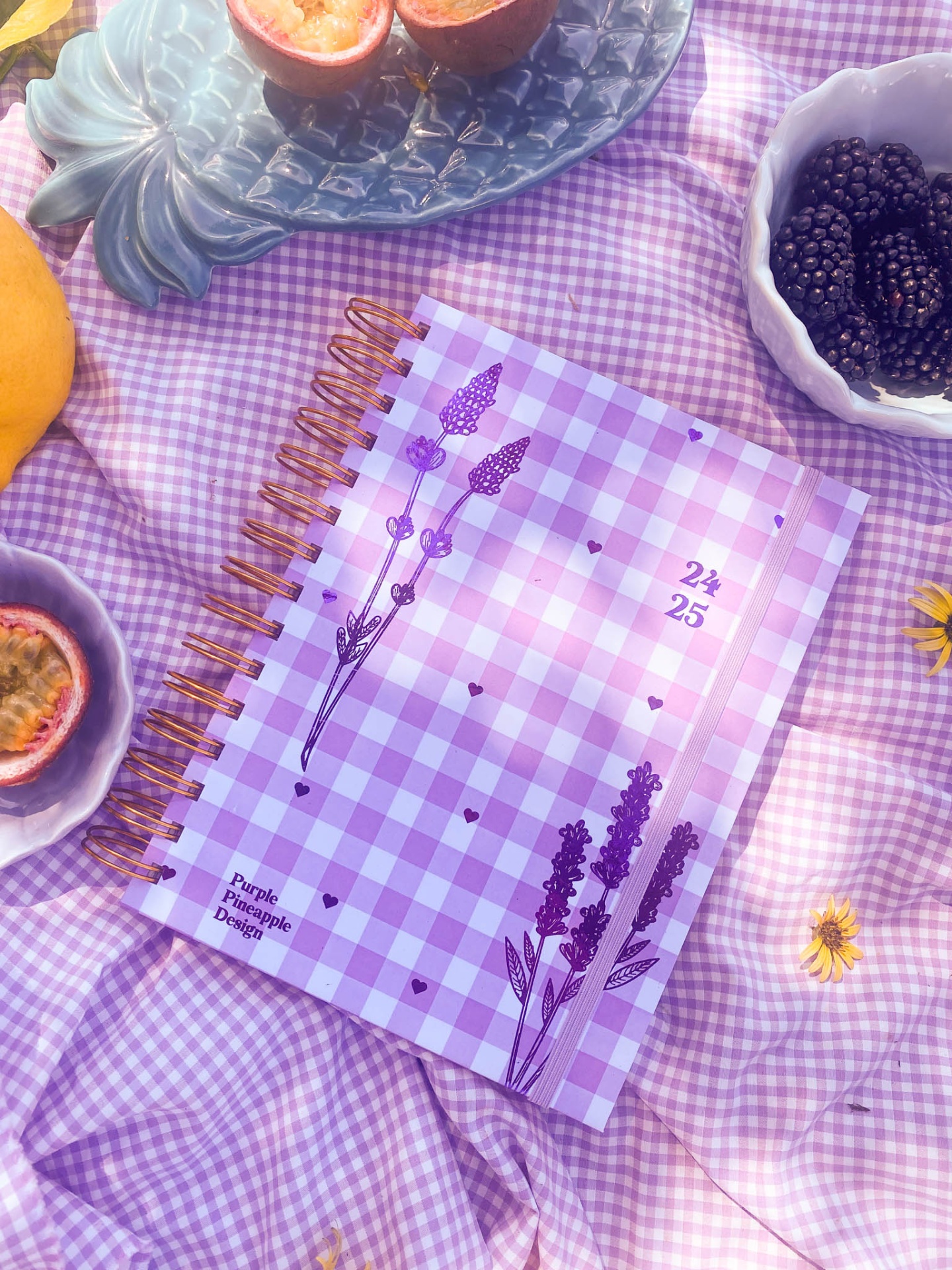 Caderno espiral lilás com flores e texto sobre tecido xadrez lilás, rodeado de fruta e pratos.