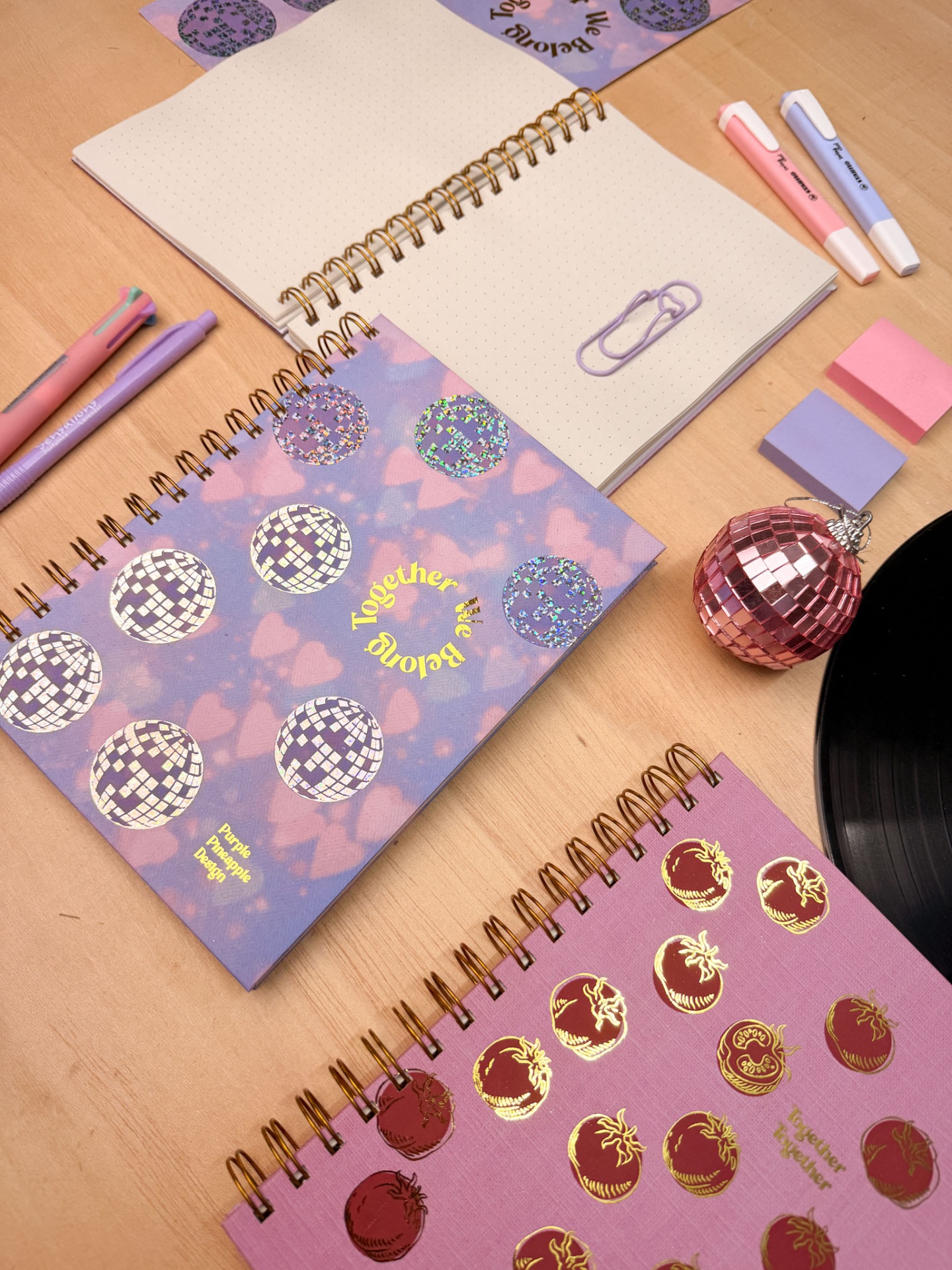 Três cadernos espiralados com estampas redondas e texto amarelo sobre mesa com canetas e blocos de notas coloridos