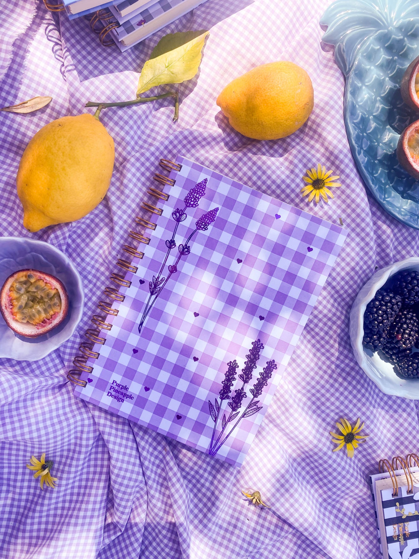 Caderno lilás quadriculado com desenhos de flores sobre tecido quadriculado lilás, rodeado por limões, amoras, maracujá e flores amarelas