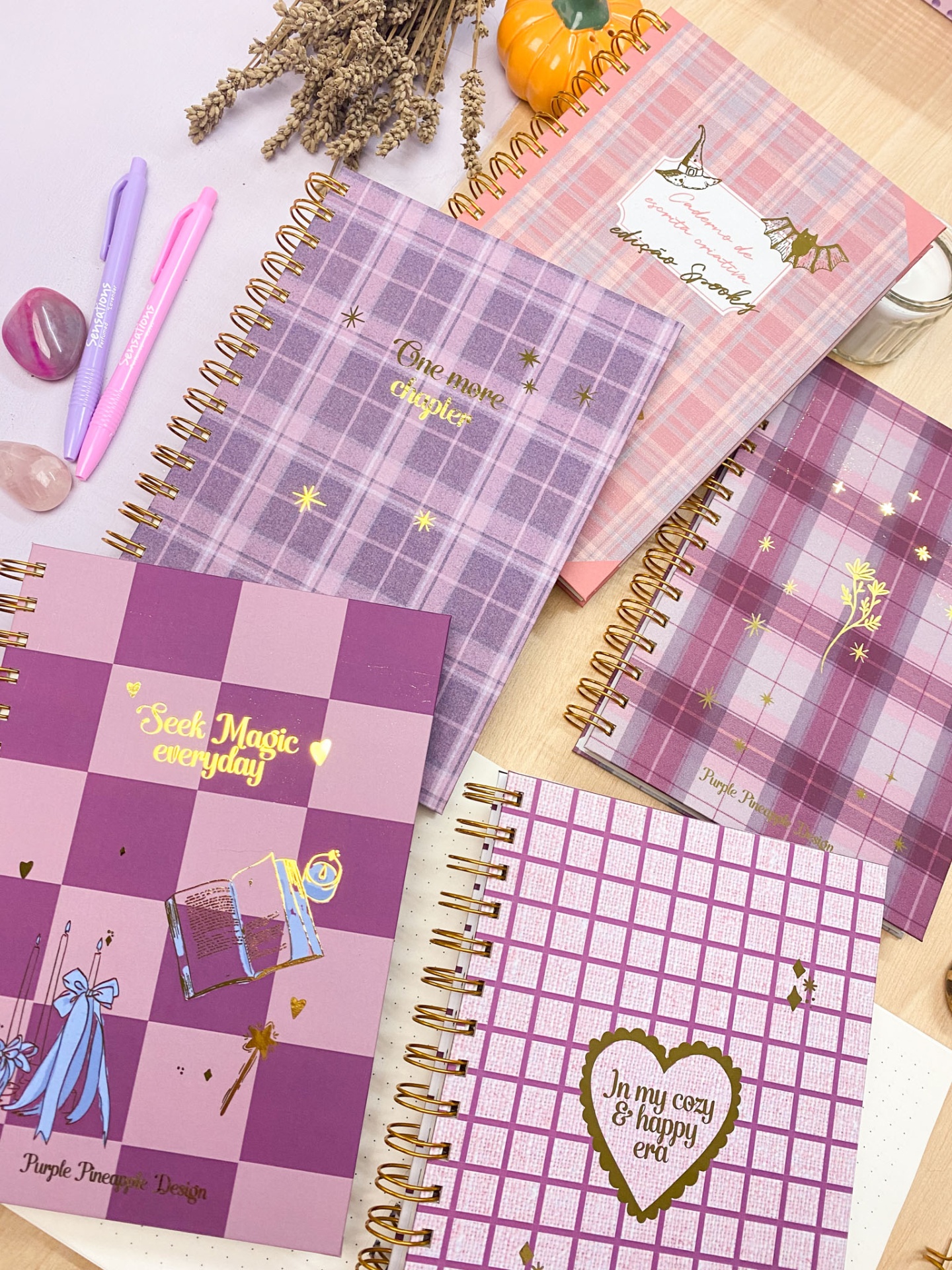 Cadernos espiral rosa e roxo com texto dourado e padrões xadrez sobre mesa com canetas e pedras