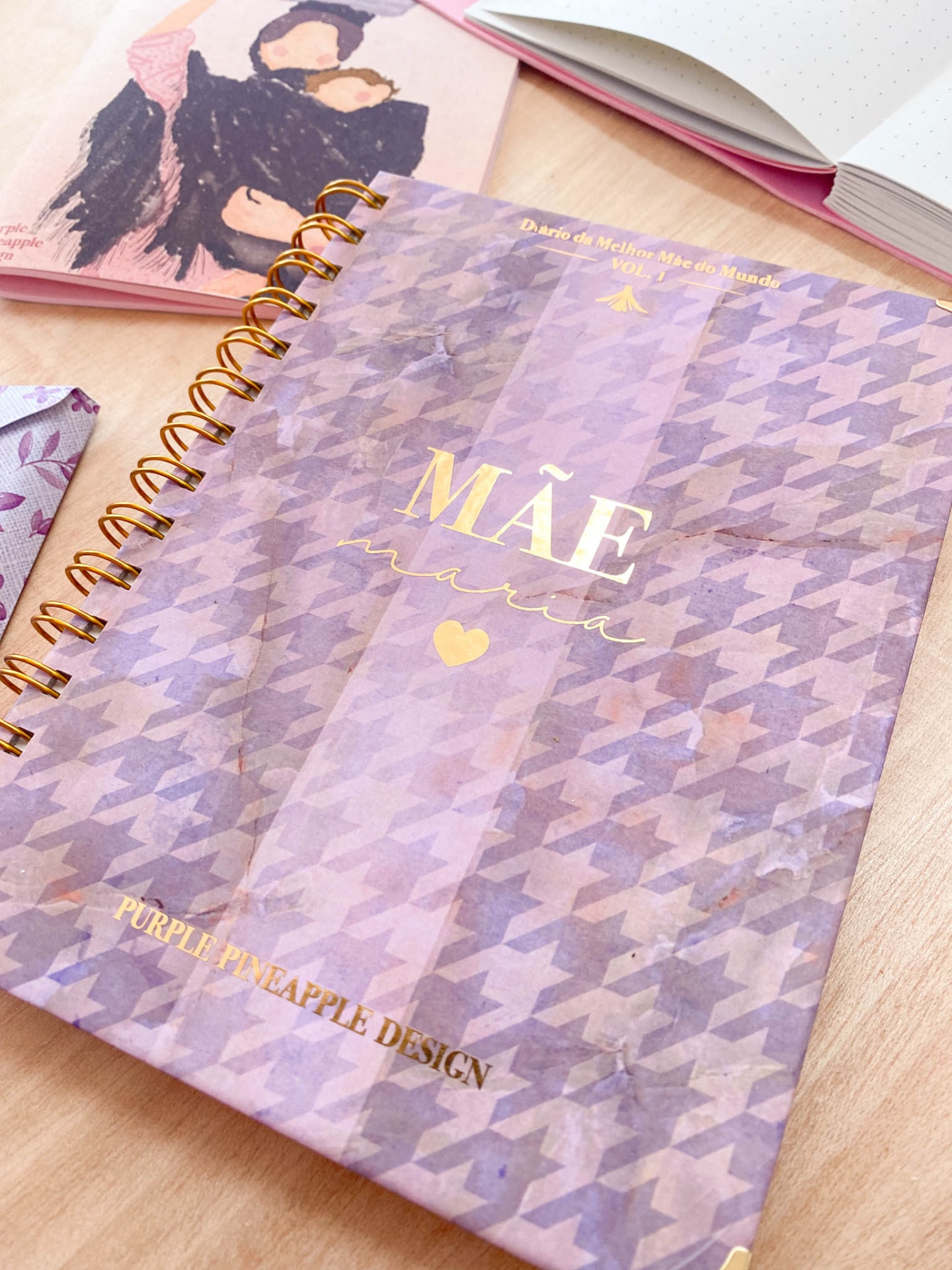 Caderno espiral lilás com padrão pied-de-poule e texto dourado 'MÃE MARAVILHA' numa mesa de madeira