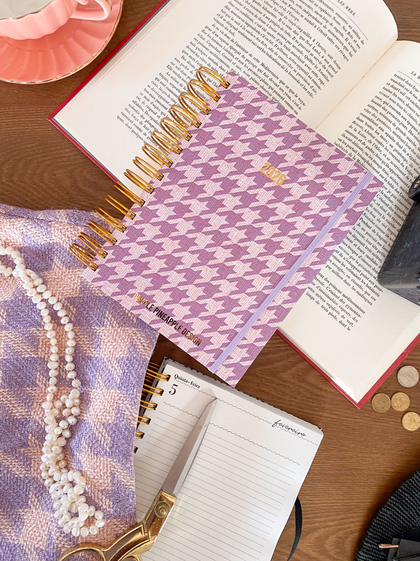 Caderno com capa pied-de-poule rosa e lilás sobre livro aberto, tecido e colar de pérolas próximo.