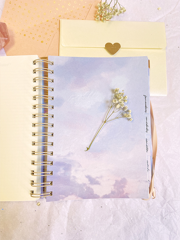 Caderno espiral aberto com capa interna de nuvens e flor secada, envelope bege com coração