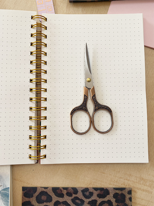 Tesoura desenhada sobre caderno pontilhado com encadernação dourada, em mesa de madeira clara