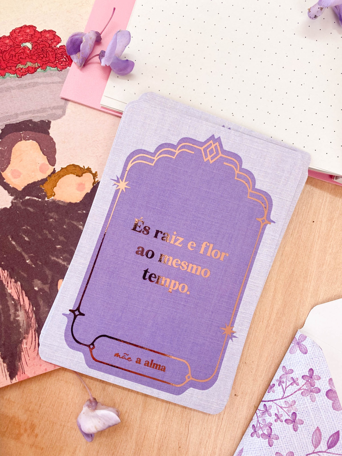 Cartão lilás com texto dourado sobre mesa de madeira com folhas e flores lilases