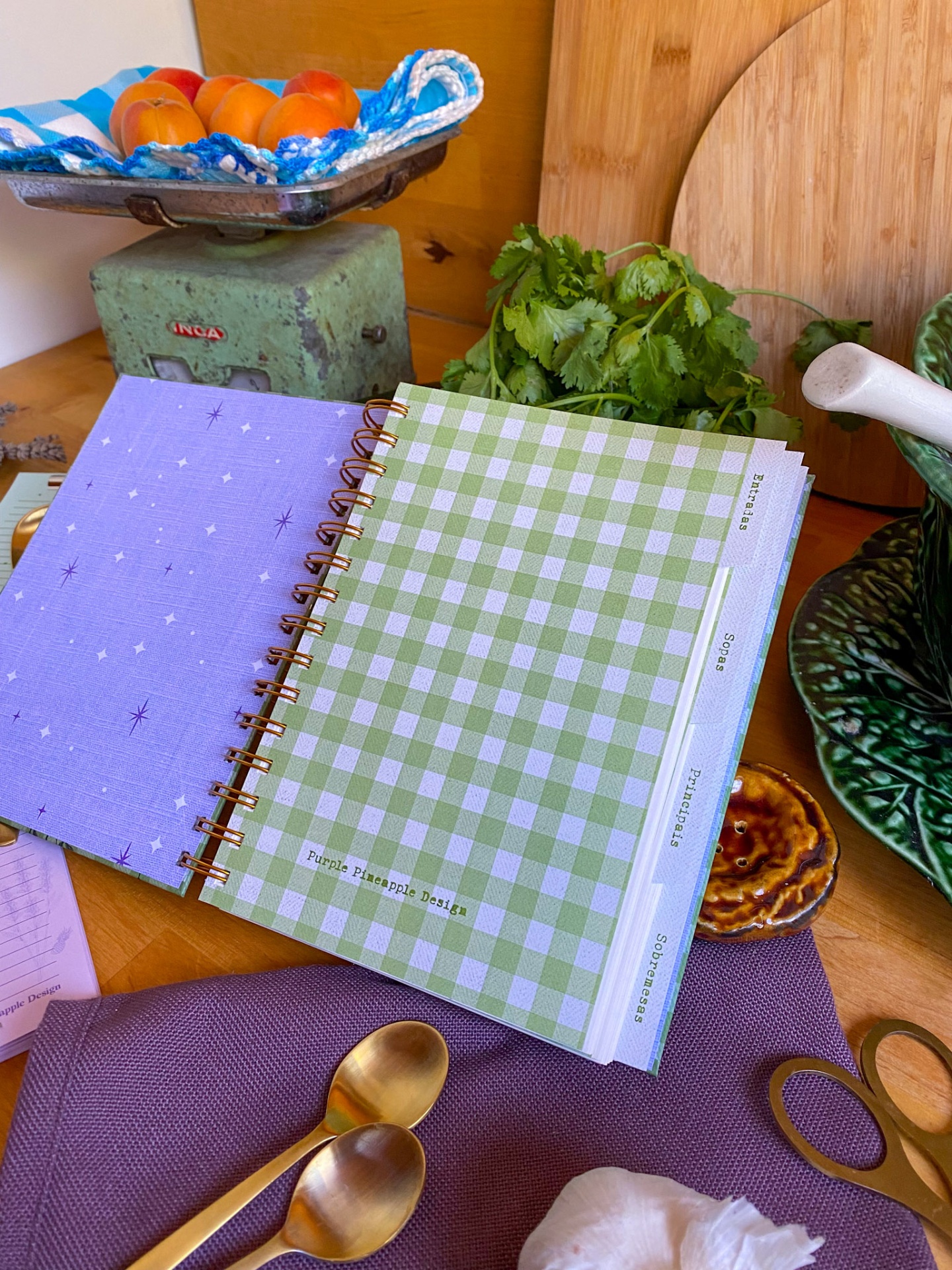 Caderno de receitas aberto com capa verde e branca às quadrículas e abas rotuladas, sobre mesa de madeira com folhas e colheres douradas.