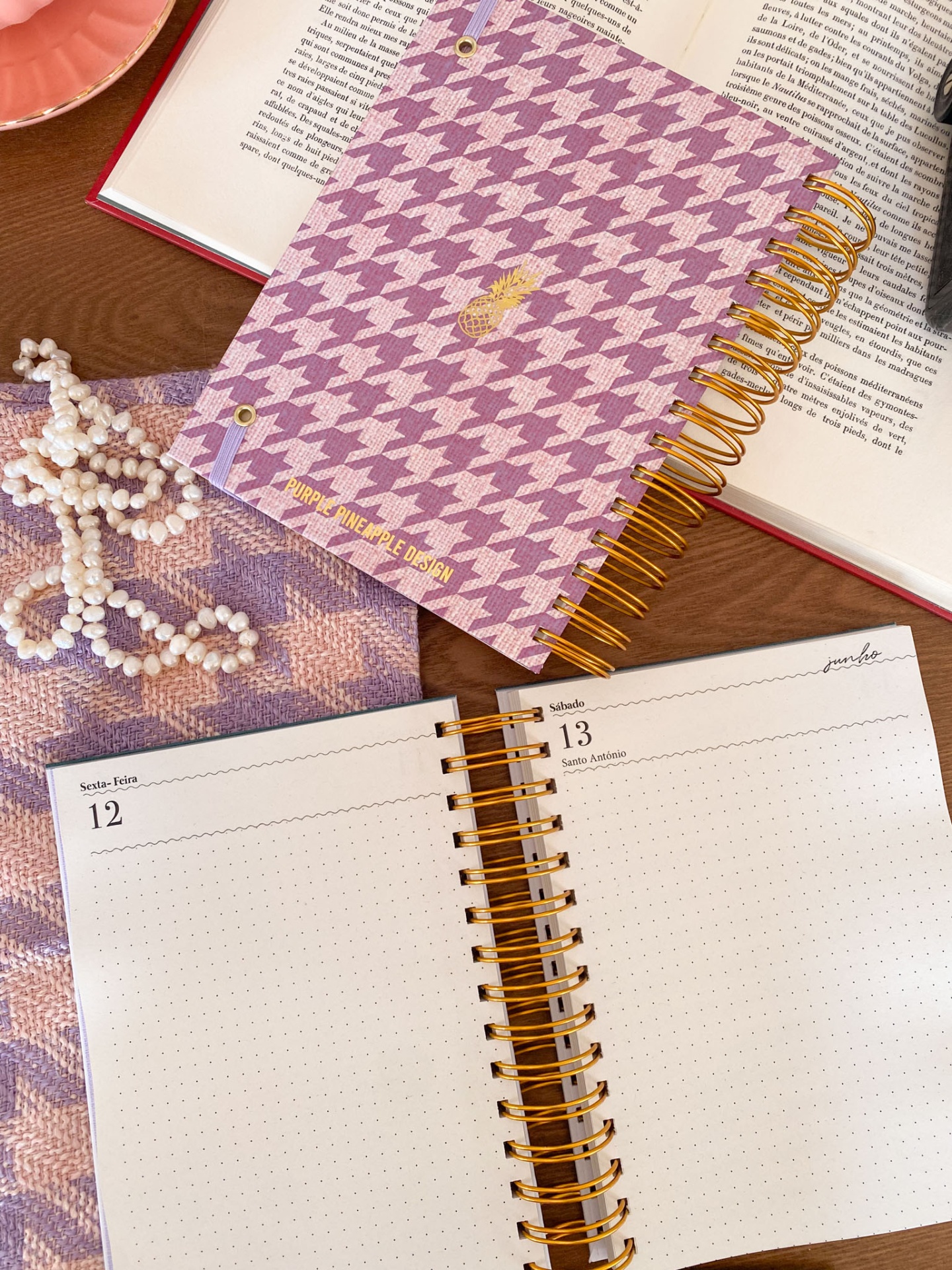 Dois cadernos espiralados com capa em padrão pied-de-poule lilás e rosa sobre mesa de madeira com pérolas e livro aberto