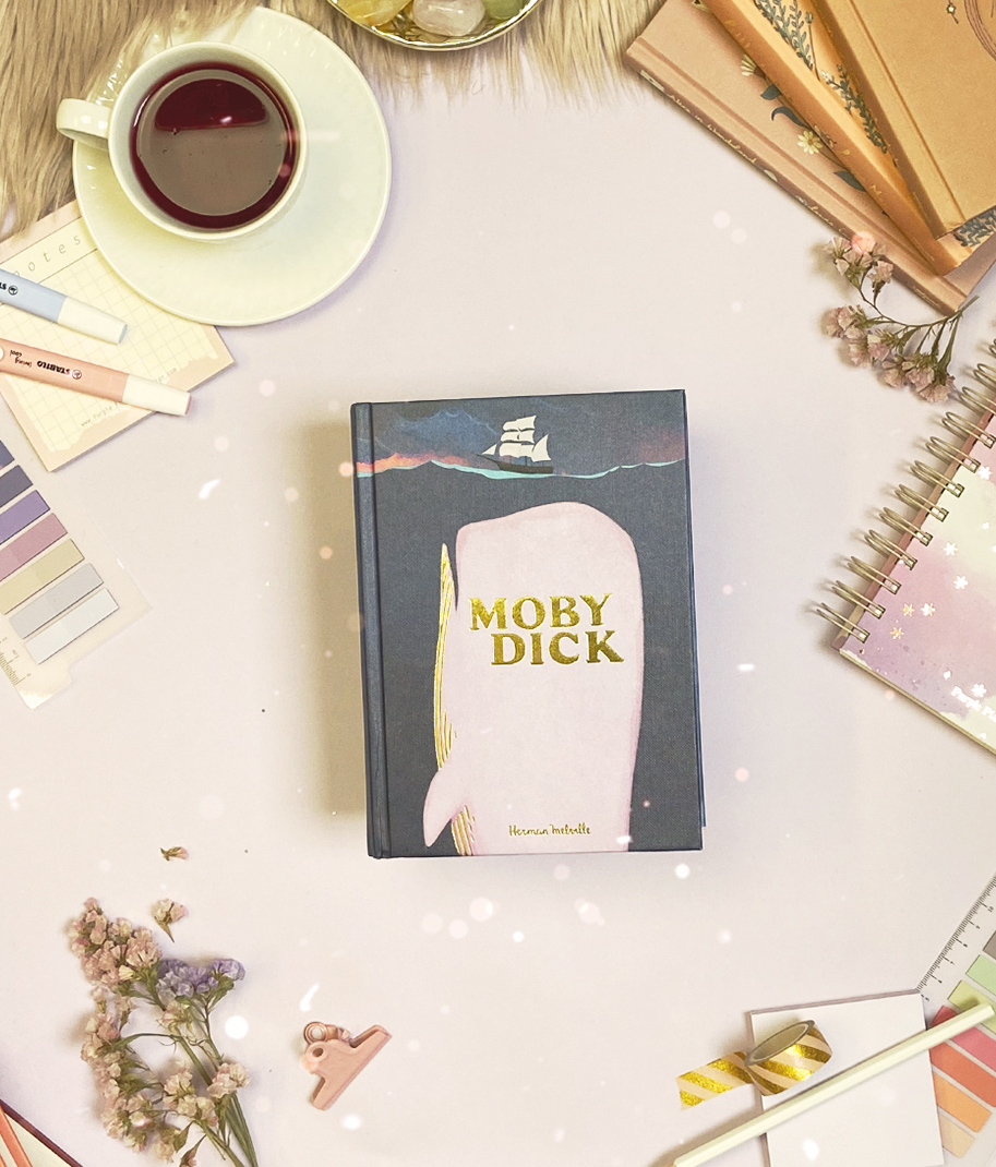 Livro Moby Dick com capa azul escuro e baleia branca numa mesa com acessórios de escritório e chá