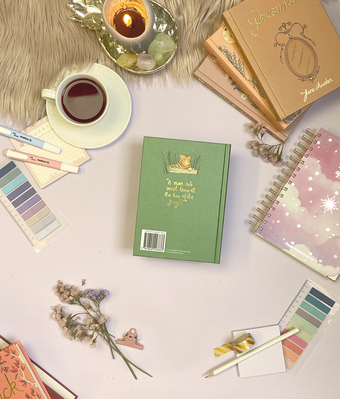 Mesa com caderno verde, caneca de chá, vela, cadernos, paletas de cor e flores secas