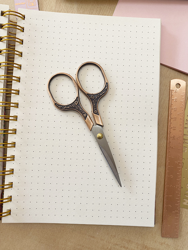 Tesoura pequena com cabo em bronze sobre caderno pontilhado com espiral dourada e régua metálica cor de cobre