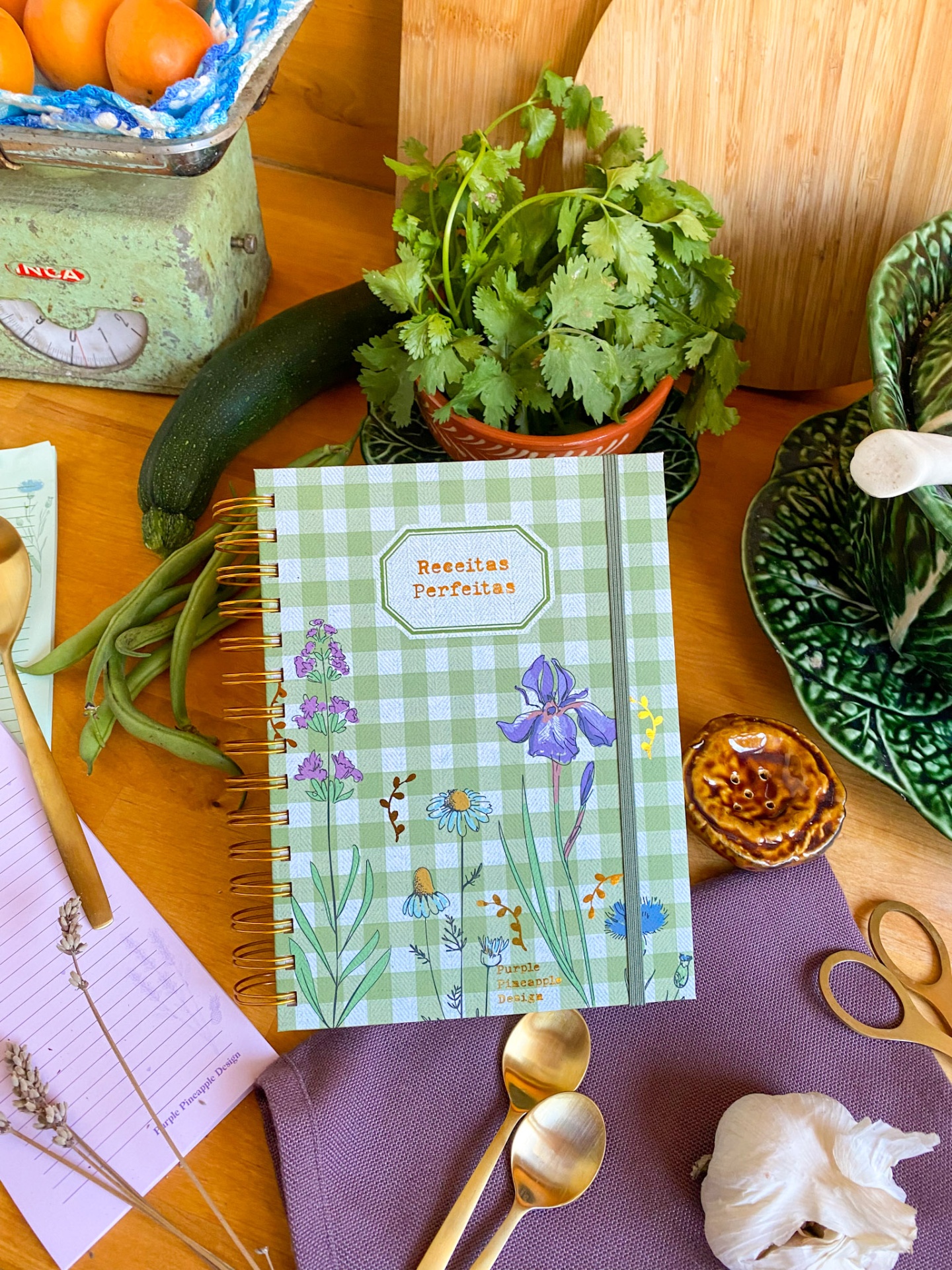 Caderno de receitas com capa verde xadrez e flores sobre mesa com legumes e utensílios