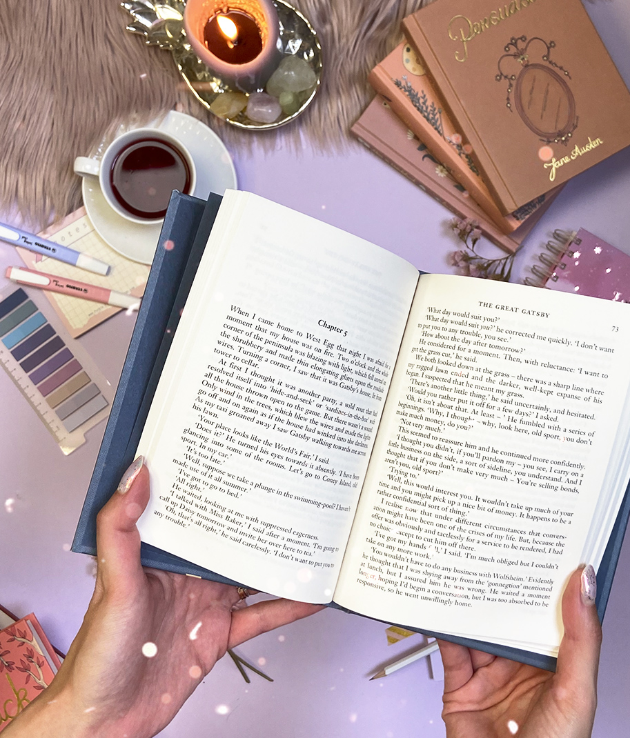 Livro aberto sendo segurado com textos em inglês, velas, café, amostras de cores, canetas e outros livros sobre superfície lilás.