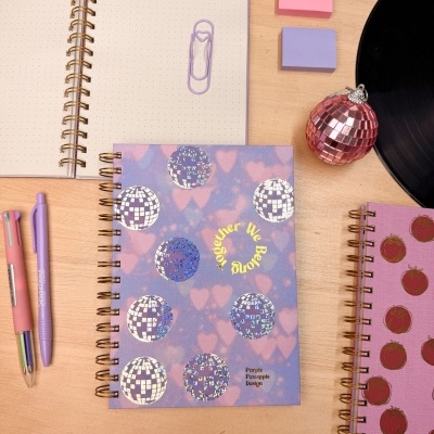 Três cadernos espirais com capas decorativas e um aberto com página pontilhada sobre mesa de madeira.