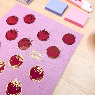 Caderno e folhas decoradas com tomates vermelhos e texto dourado, blocos de notas rosa e roxo e marcadores numa mesa de madeira.