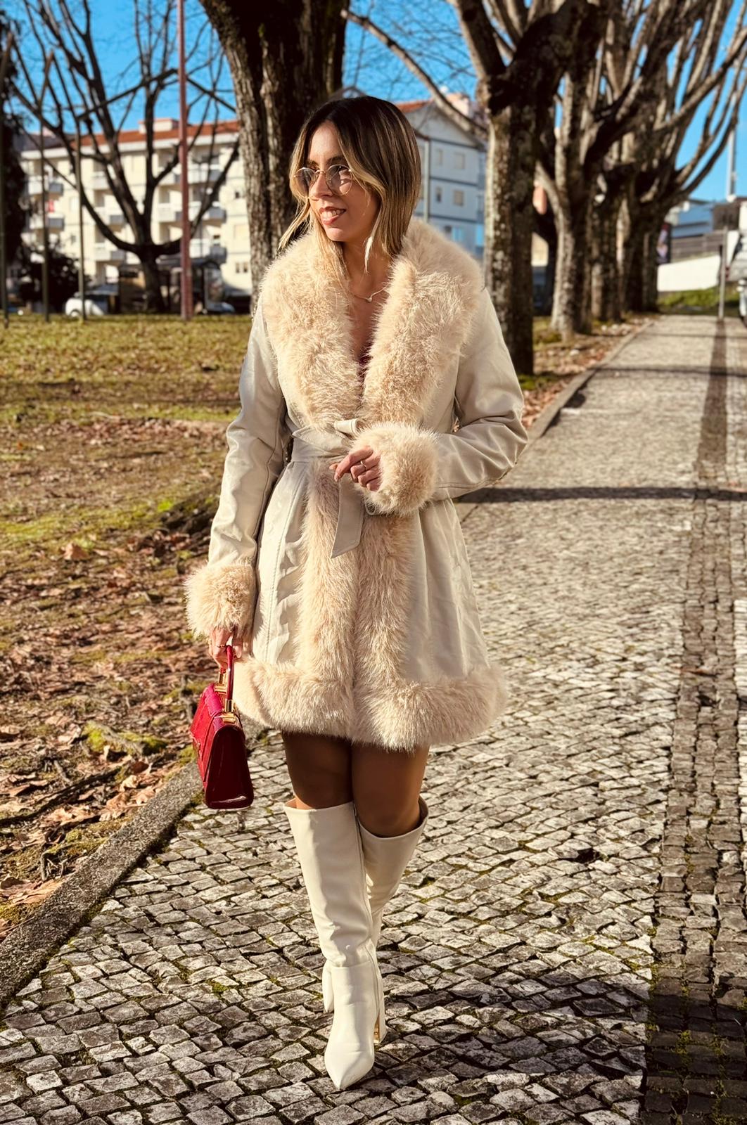 Casaco Napa c/ Pêlo Mulher com casaco bege de pelo, botas brancas e mala vermelha a caminhar em passeio com calçada e árvores.