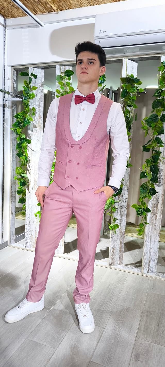 Fato Rosa Ponteado Homem de fato cor-de-rosa, camisa branca e gravata borboleta vermelha em ambiente interior decorado com madeira branca e plantas verdes.