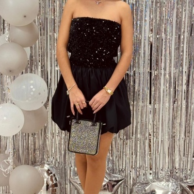 Mulher com vestido curto preto de lantejoulas, bolsa pequena e sapatos brilhantes, em ambiente decorado com balões prateados e brancos.