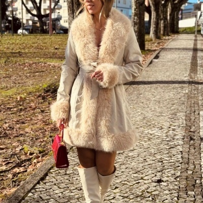 Mulher com casaco bege de pelo, botas brancas e mala vermelha a caminhar em passeio com calçada e árvores.