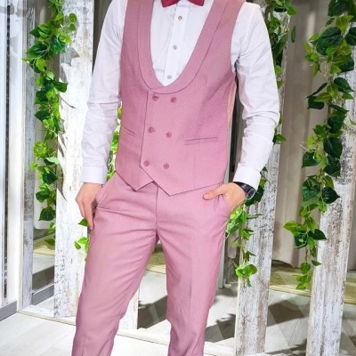 Homem de fato cor-de-rosa, camisa branca e gravata borboleta vermelha em ambiente interior decorado com madeira branca e plantas verdes.