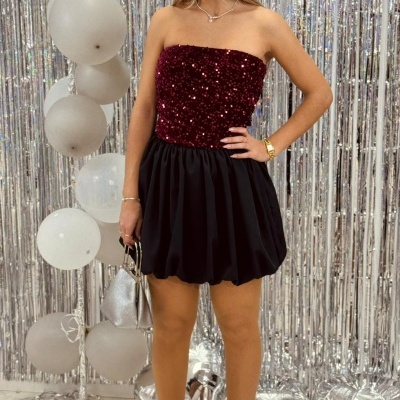 Mulher com vestido sem alças bordô e preto, sapatos e mala prateados, fundo com cortina e balões prateados