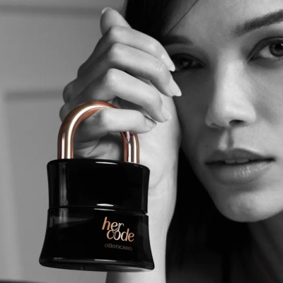 Frasco em forma de cadeado preto com tampa cobre 'her code O BOTICÁRIO' segurado pela mão de mulher