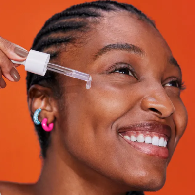 Mulher com sérum facial em conta-gotas e brincos coloridos, fundo laranja