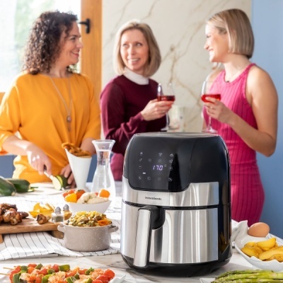 Frigideira elétrica air fryer inox e preta com visor digital na mesa com comida e três mulheres ao fundo.
