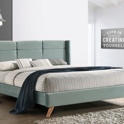 Cama estofada azul claro com cabeceira almofadada, colcha bege, manta azul escura e quadro com texto motivacional na parede