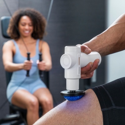 Dispositivo de massagem muscular branco com luz azul em perna, com mulher a treinar ao fundo