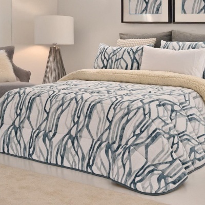 Quarto com cama vestida com edredão e almofadas de padrão abstrato azul e cinza, cobertor de pelo bege, cadeira cinza e candeeiro