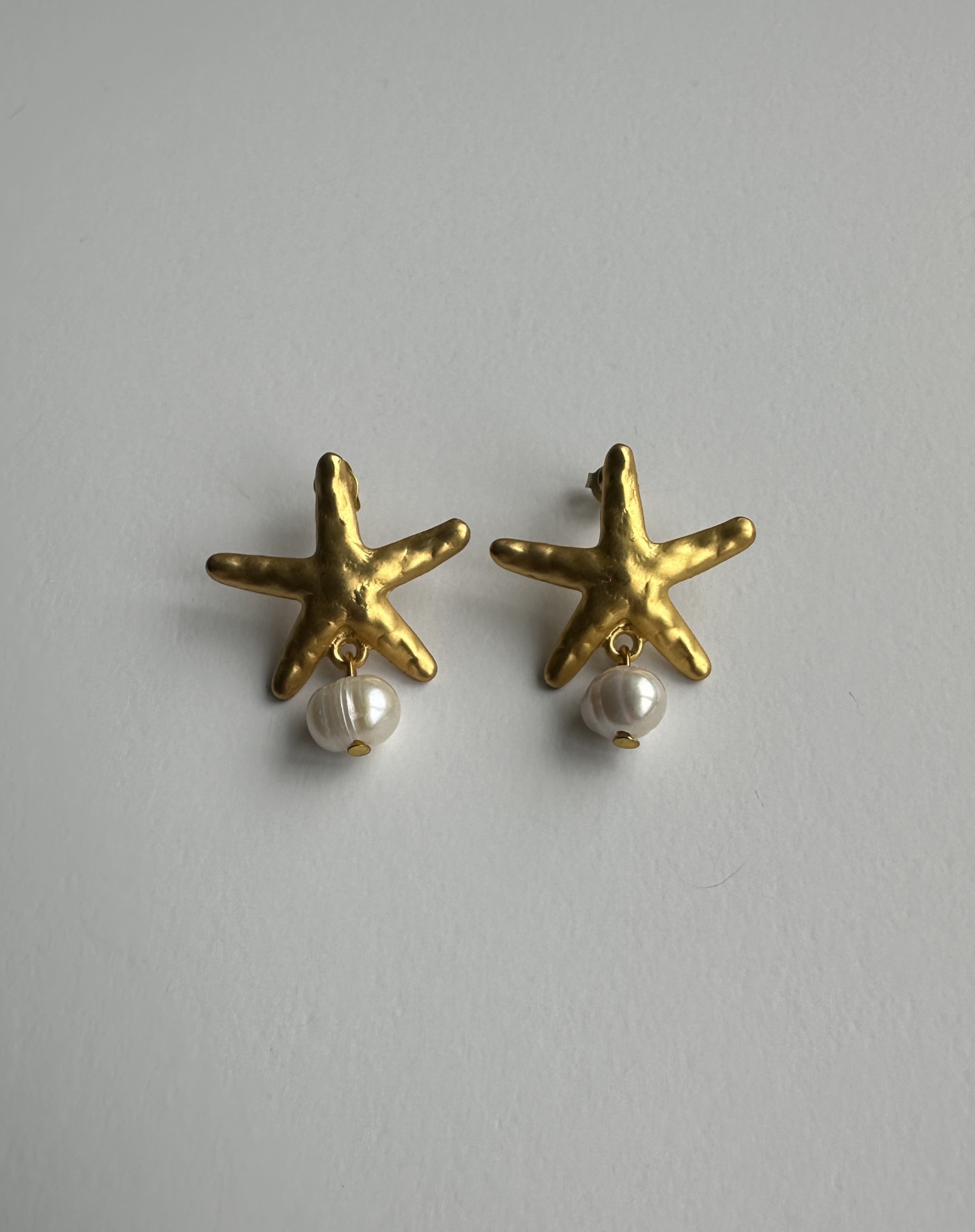 Brincos Estrela-do-Mar Dourados com Pérola – Toque Náutico Sofisticado para Todo o Ano Brincos dourados em forma de estrela do mar com pérola pendente.