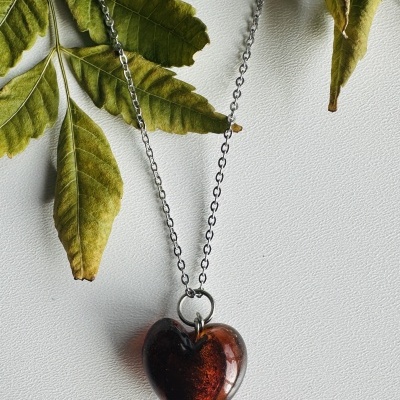 Colar com coração de vidro vermelho e corrente prateada em fundo branco com folhas verdes.