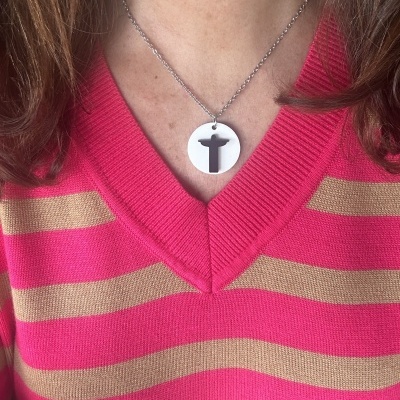 Colar com pendente redondo prateado com recorte Cristo Redentor, usado com camisola de riscas rosa e bege.