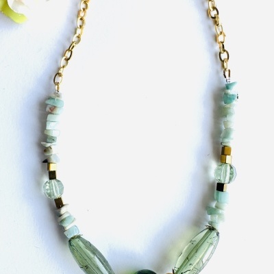 Colar dourado com contas verdes e azuis sobre fundo branco com flor cor-de-rosa