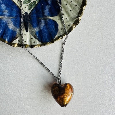 Colar com coração dourado e castanho em corrente de metal prateado sobre tecido com borboleta azul