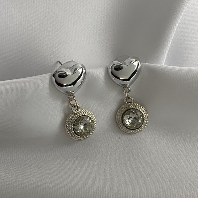 Brincos prateados em forma de coração com pendente de pedra transparente e aro dourado, sobre tecido branco.