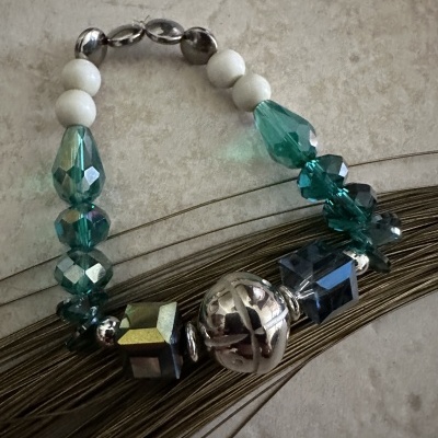 Pulseira com contas verdes, azuis, brancas e metálicas sobre fundo claro e linhas castanhas