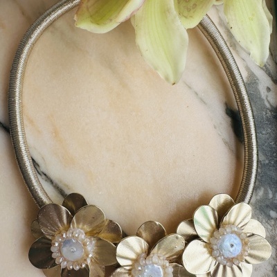 Colar dourado com três flores decorativas com pedras brancas, sobre mármore e flores naturais.