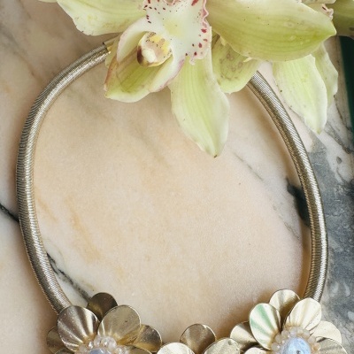 Colar dourado com flores metálicas e pedras brancas junto a flores naturais verdes e brancas