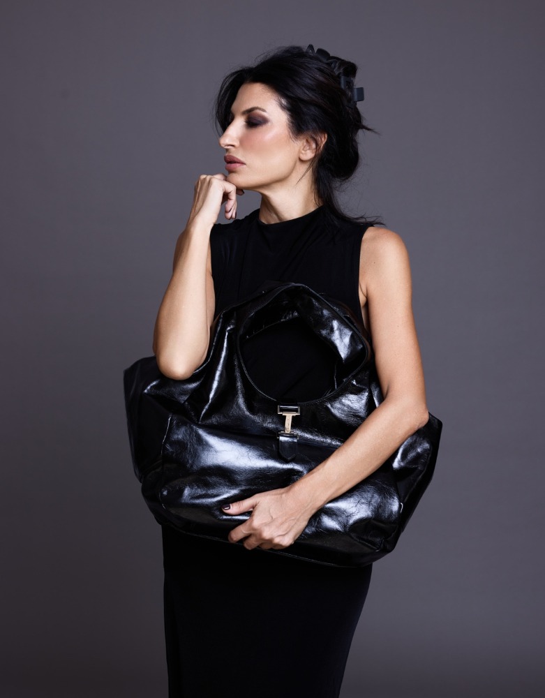 Mulher com vestido preto segurando mala oversized preta de material brilhante com fecho de metal prateado.
