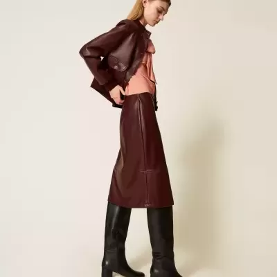 Mulher a usar saia midi e casaco castanhos escuros de couro, camisa rosa clara e botas pretas até ao joelho, em fundo neutro.