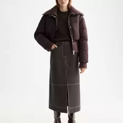 Mulher com casaco acolchoado castanho escuro, saia comprida castanha com costuras brancas e botas castanhas