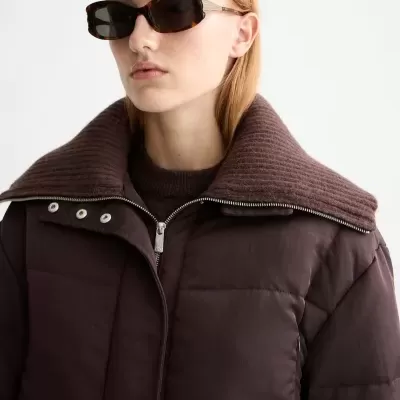 Casaco acolchoado castanho escuro com gola alta de malha e óculos escuros.
