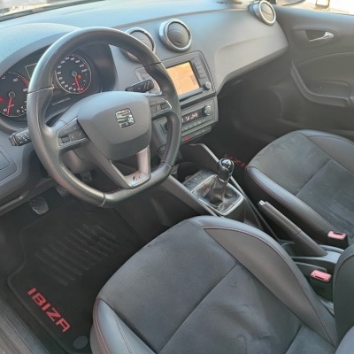 Interior de carro SEAT Ibiza com volante em couro, bancos cinza e preto, painel cinza e tapete preto com texto vermelho