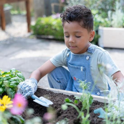 Criança a cuidar de plantas num canteiro de jardim com enxada e luvas.